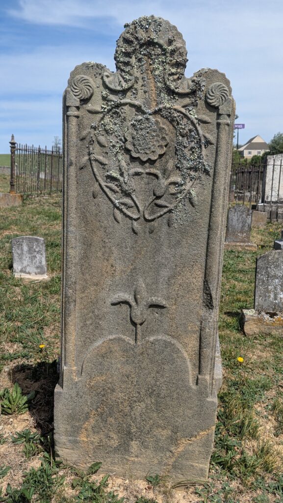 Tall granite headstone carved by Laurence Krone with an ornate sunflower-and-vine wreath enclosing a rose bloom, lily-heart motif beneath